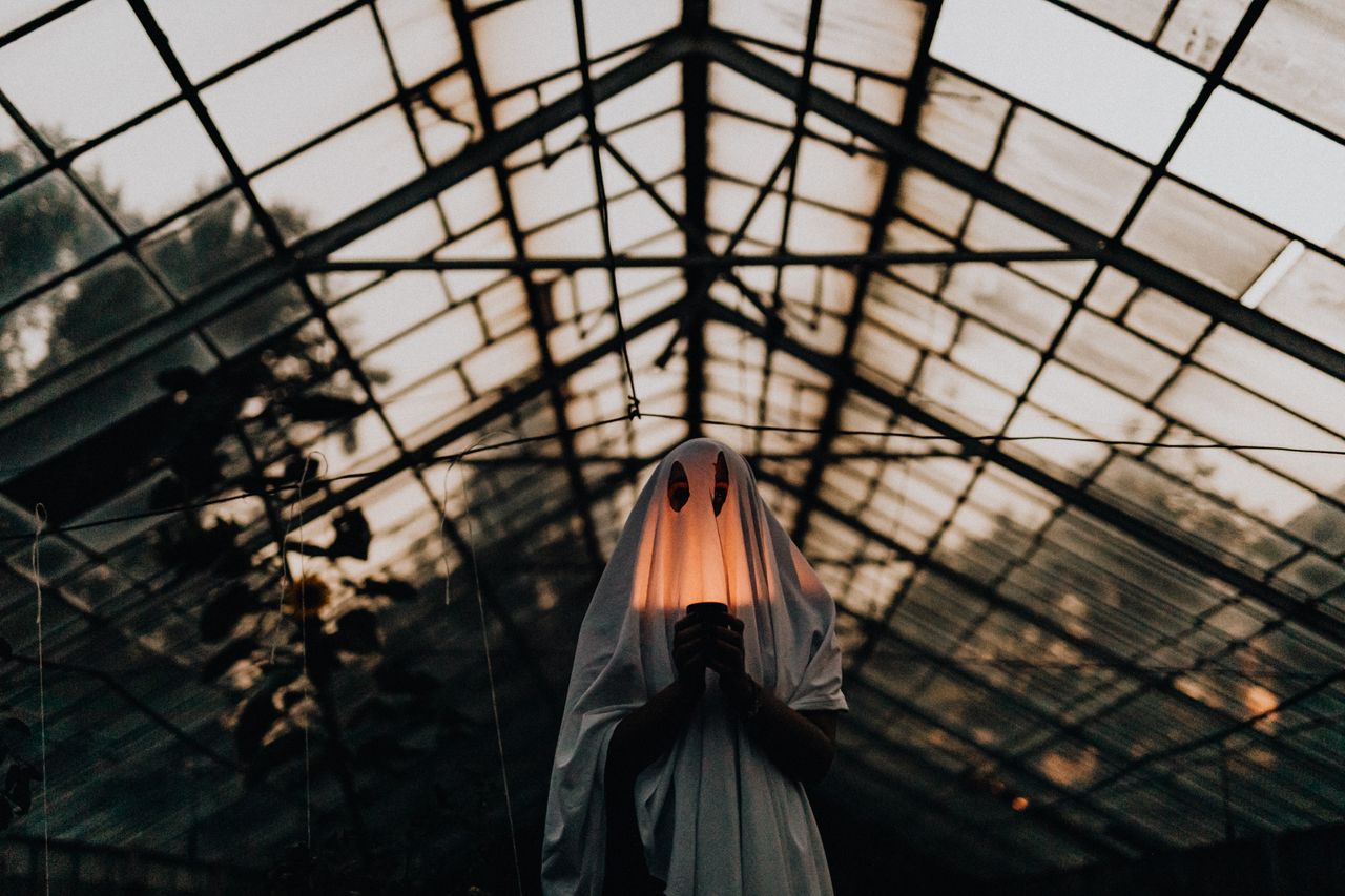 Services Mysterious figure in ghost costume holding lantern inside a dimly lit greenhouse.